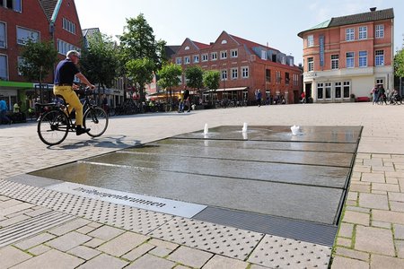 [Translate to Französisch:] Die großformatigen Natursteinplatten des Brunnenareals sind minimal gestuft, um ein Gefälle zu erzeugen. Dennoch ist die Fläche völlig barrierefrei und kann selbst von Radfahrern durchquert werden.