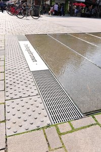 Im unteren Bereich bildet eine Stabile-Rinne mit Längsstabrosten den Abschluss. Auf eine mittig platzierte Edelstahlplatte wurde vom Metallwarenhersteller der Schriftzug „Dreiburgenbrunnen“ eingelasert.